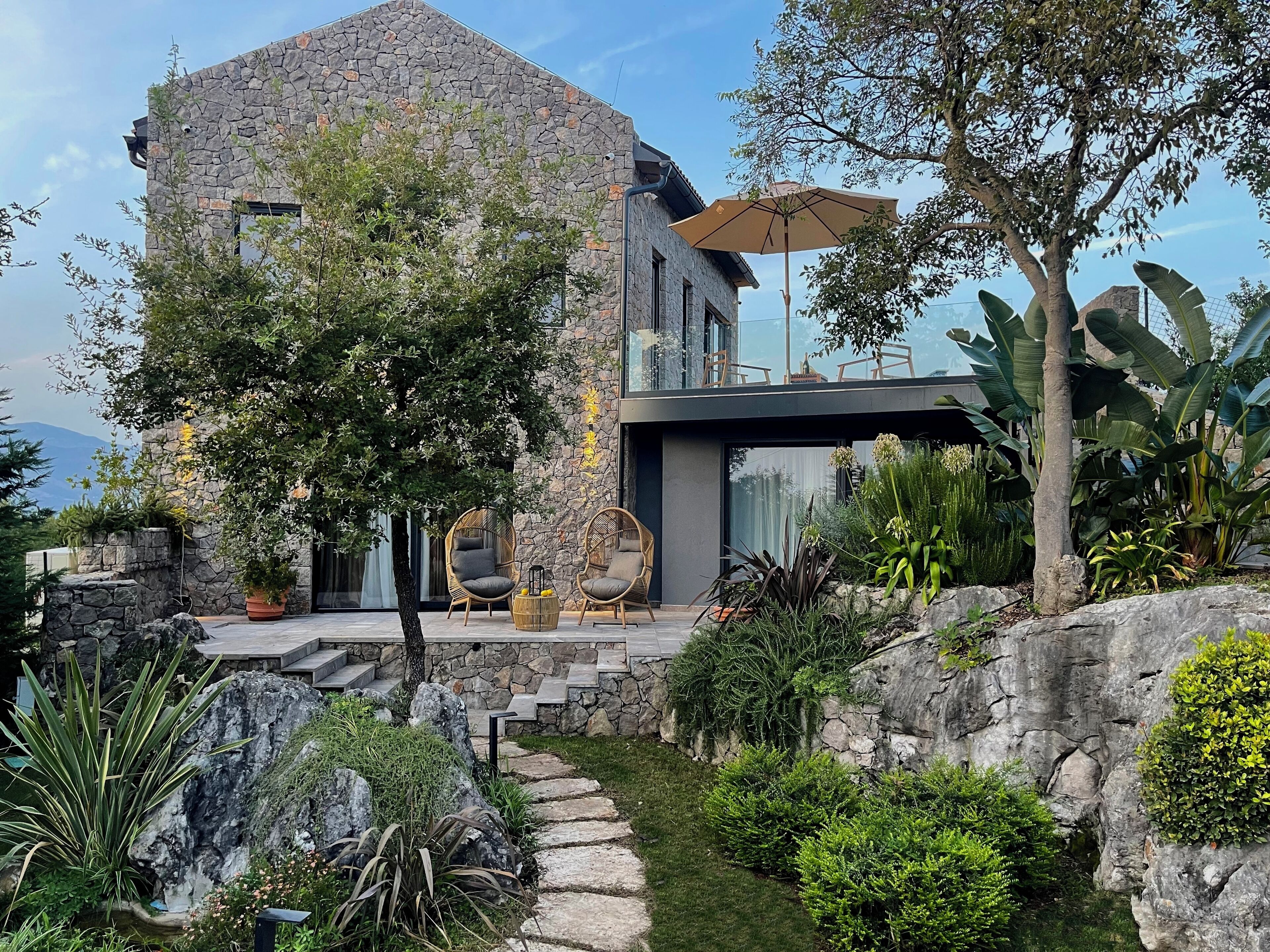 Una casa moderna con fachada de piedra, un balcón con sillas de mimbre y un jardín con diversas plantas y un sendero de piedra.