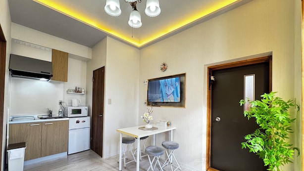 Uma cozinha compacta com eletrodomésticos modernos, uma área de refeições com mesa e banquetas, uma televisão fixada na parede, e uma porta com um vaso de planta ao lado.