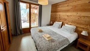 A bedroom with a wooden headboard, a large bed with a patterned bedspread, two bedside tables with lamps, and a view of a wooden deck through a glass door. - L'Echaillon - Soldanelle (Névache)