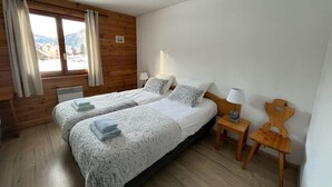 Quarto com duas camas, paredes de madeira, janela com cortinas, mesa de cabeceira com luminária e uma cadeira.