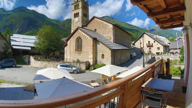 Ein Holzbalkon mit Blick auf ein Dorf, eine Kirche, einen Berg im Hintergrund und geparkte Autos.