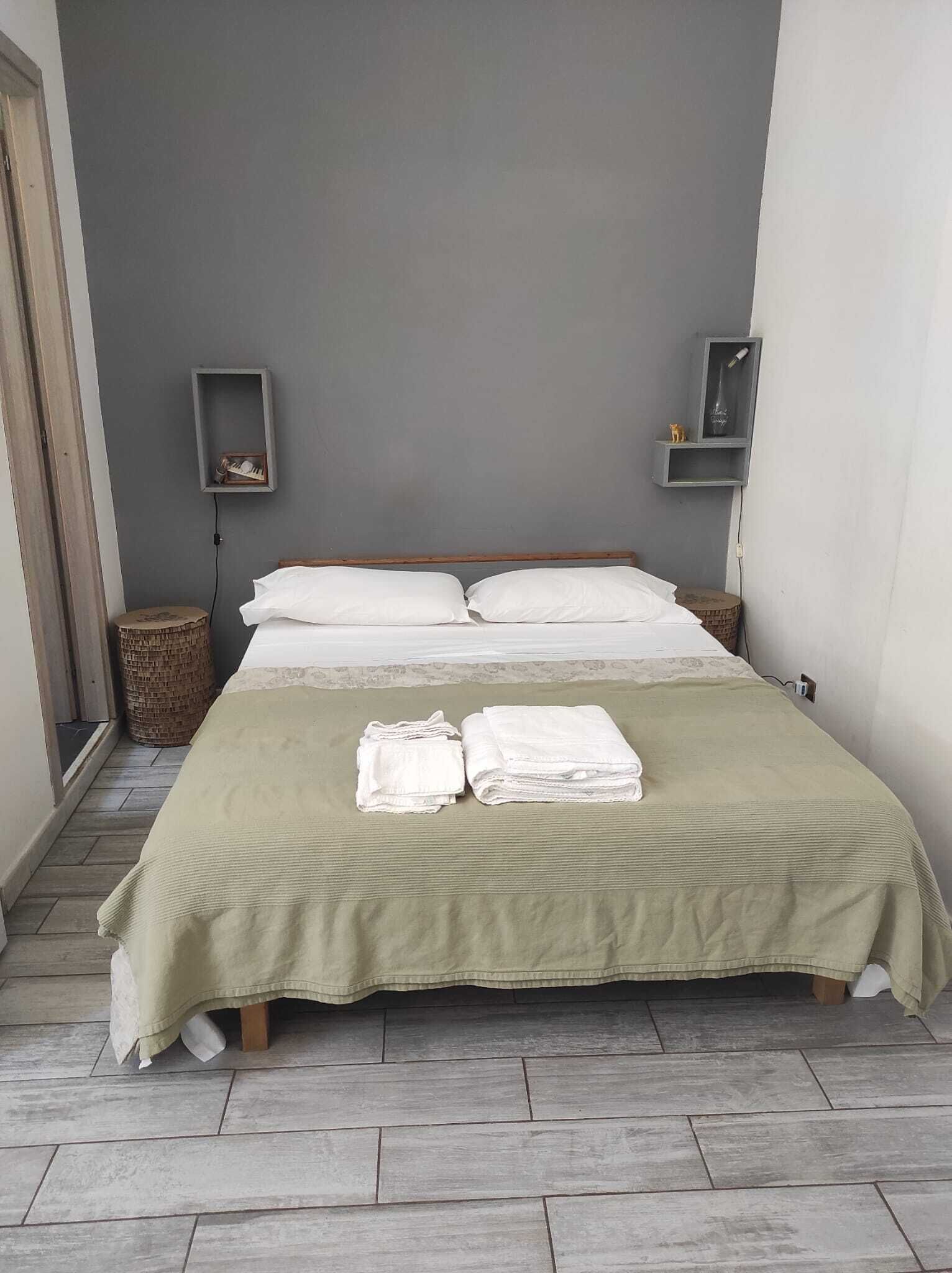 Ein ordentlich bezogenes Bett mit grüner Decke und weißen Kissen, zwei Nachttische mit kleinen Ablagen und ein Korbwäschekorb auf dem Boden.