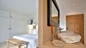 Un dormitorio con una cama, un banco de madera, un espejo grande y un baño con un lavabo y una puerta de madera.