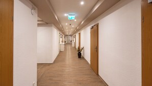 Un long couloir étroit, doté d’un plancher en bois et de murs blancs. Des portes se trouvent de part et d’autre, et une plante en pot est visible au centre.