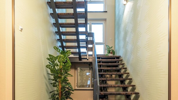 Een houten trap met een metalen leuning, een potplant en een muur met een reliëfpatroon.