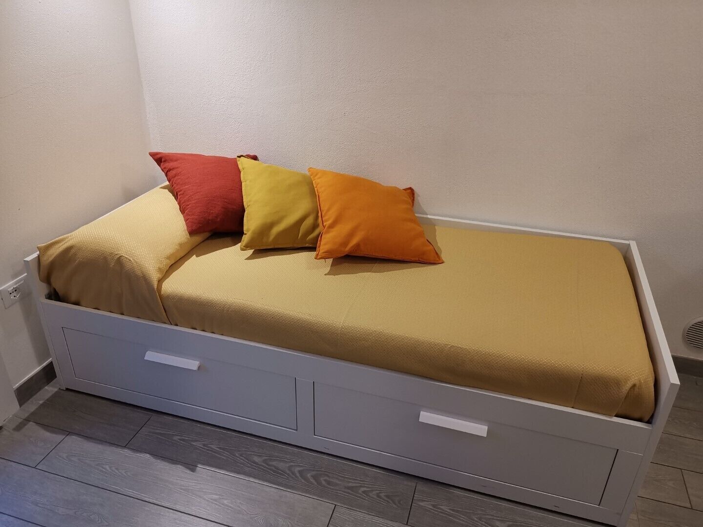 A small bed with a beige mattress and three pillows in red, yellow, and orange.