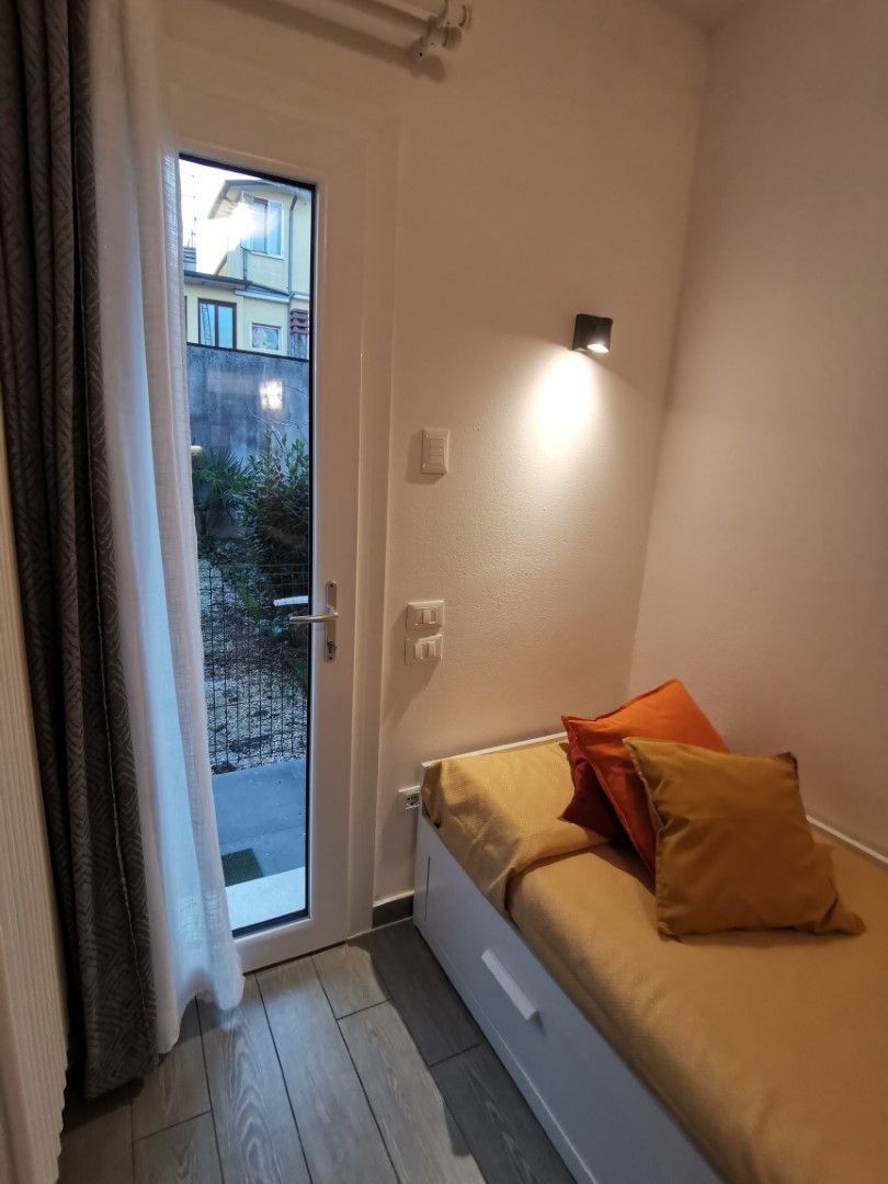Una stanza piccola e compatta con un letto singolo, una porta scorrevole su un balcone e una piccola luce fissata al muro.