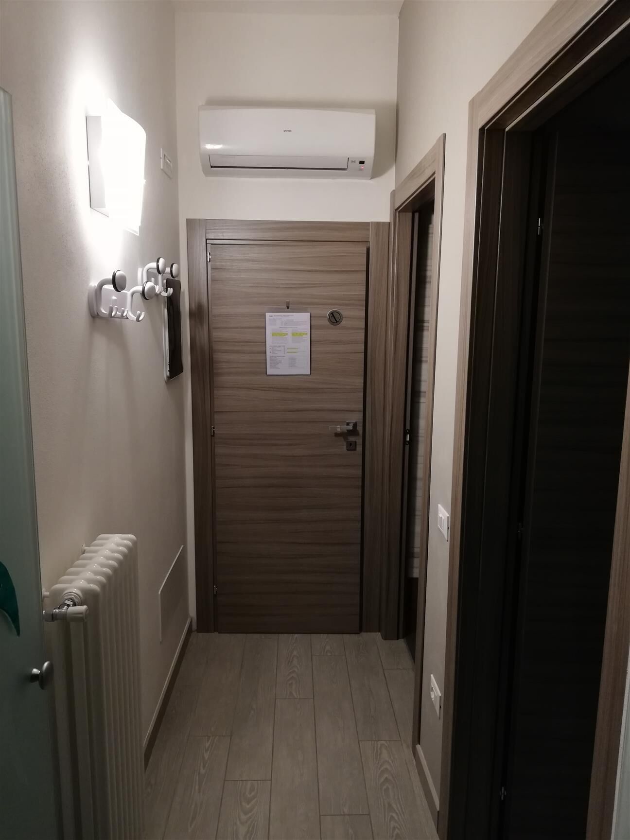 A hallway with a radiator, a wall-mounted air conditioner, a door with a notice, and a coat rack.