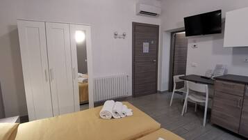 A hotel room with a bed, a desk, a chair, a wardrobe, an air conditioning unit, and a television.