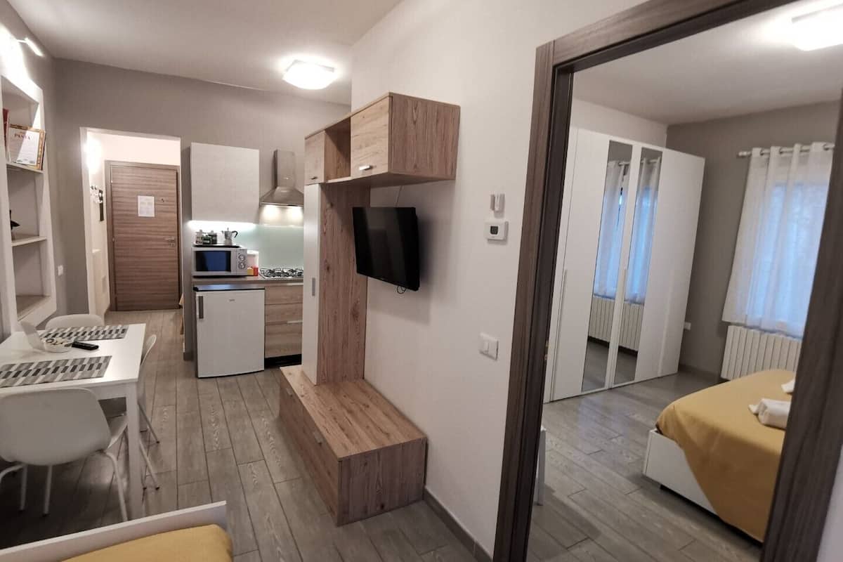Ein kompaktes Hotelzimmer mit Küchenzeile, einem an der Wand befestigten Fernseher und einem Bett mit gelber Tagesdecke.