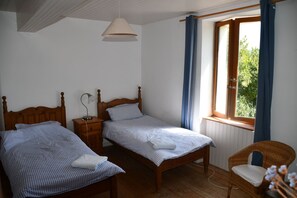 Une chambre avec deux lits simples, une commode en bois, une chaise, une fenêtre avec des rideaux bleus et une lampe suspendue.