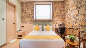 Une chambre avec un mur en pierre, un lit avec une literie jaune, un bureau avec une chaise et une petite table avec une plante.