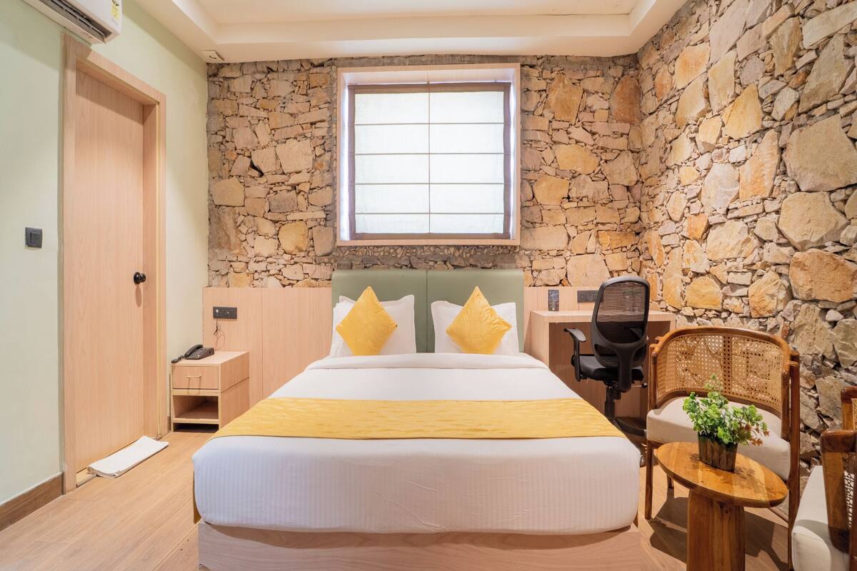 Une chambre avec un mur en pierre, un lit avec une literie jaune, un bureau avec une chaise et une petite table avec une plante.