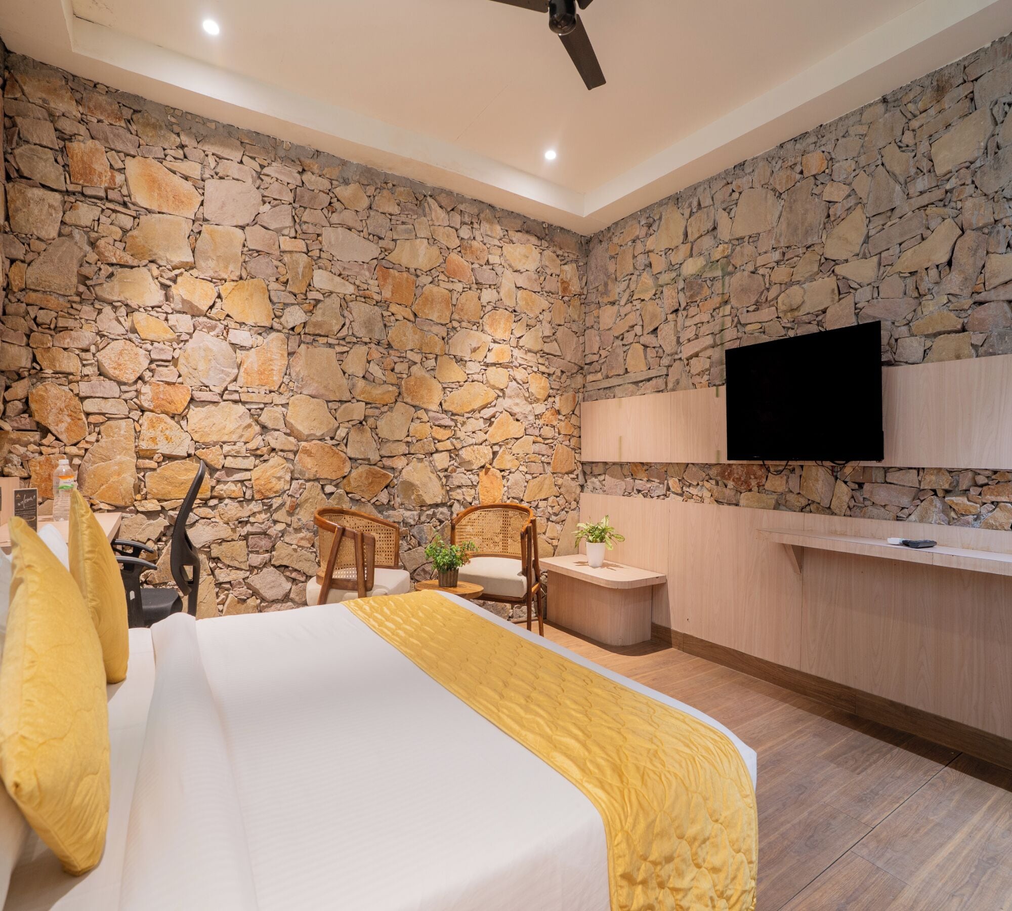 Una habitación de hotel moderna con una pared de piedra, una cama con ropa blanca y un plaid amarillo, un escritorio de madera y un televisor de pantalla plana montado en la pared.