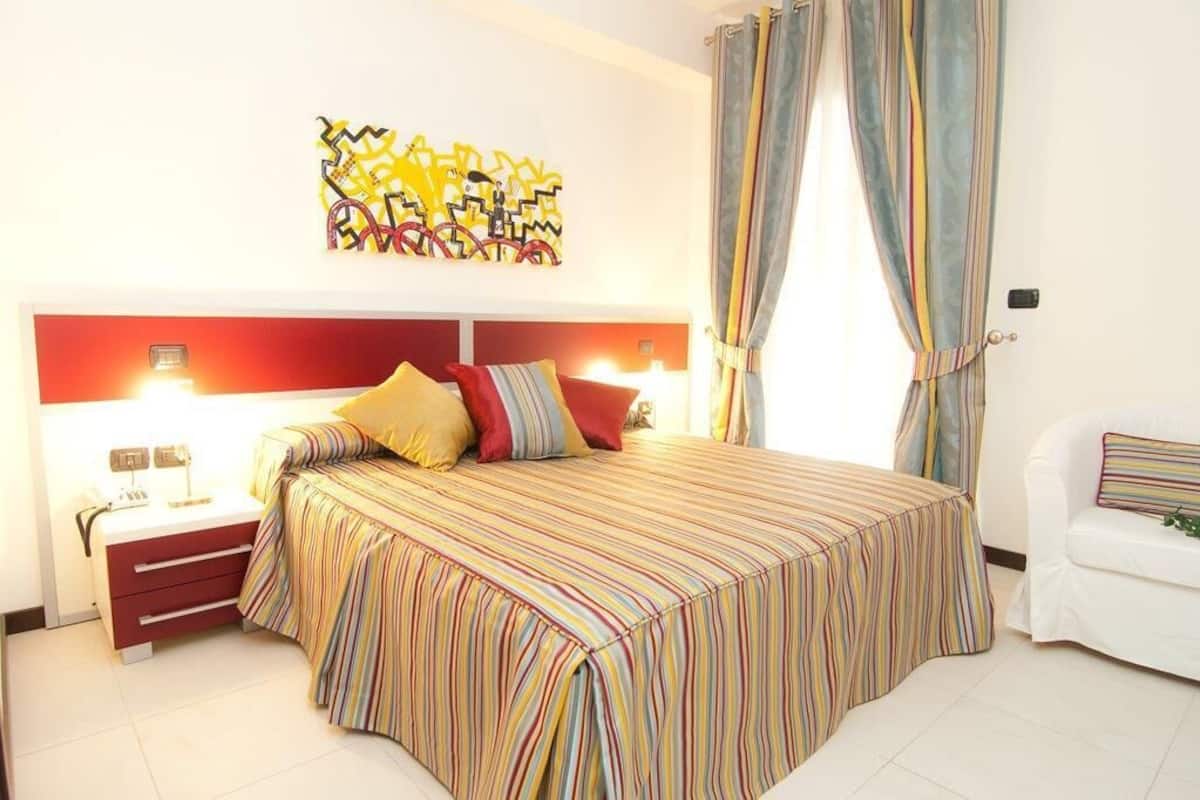 Une chambre d’hôtel avec un lit, des tables de chevet, une petite table, un canapé blanc et un tableau abstrait aux couleurs vives accroché au mur.