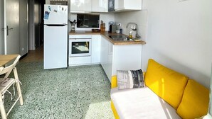 Cocina compacta con gabinetes blancos, microondas y refrigerador. Hay una zona de comedor con una mesa y sillas de madera. Un sofá amarillo está ubicado cerca de la ventana y hay un mapa colgado en la pared.