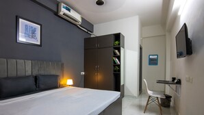 Ein modernes Hotelzimmer mit einem großen Bett, einem Kleiderschrank, einem Schreibtisch mit Stuhl, einem an der Wand montierten Fernseher und einem gerahmten Bild.