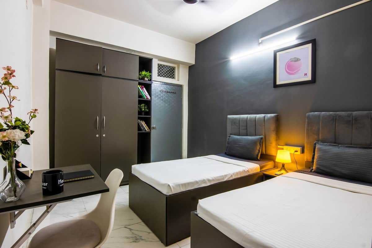 A modern hotel room with two single beds, a desk with a chair, a wardrobe, and a bedside table with a lamp.