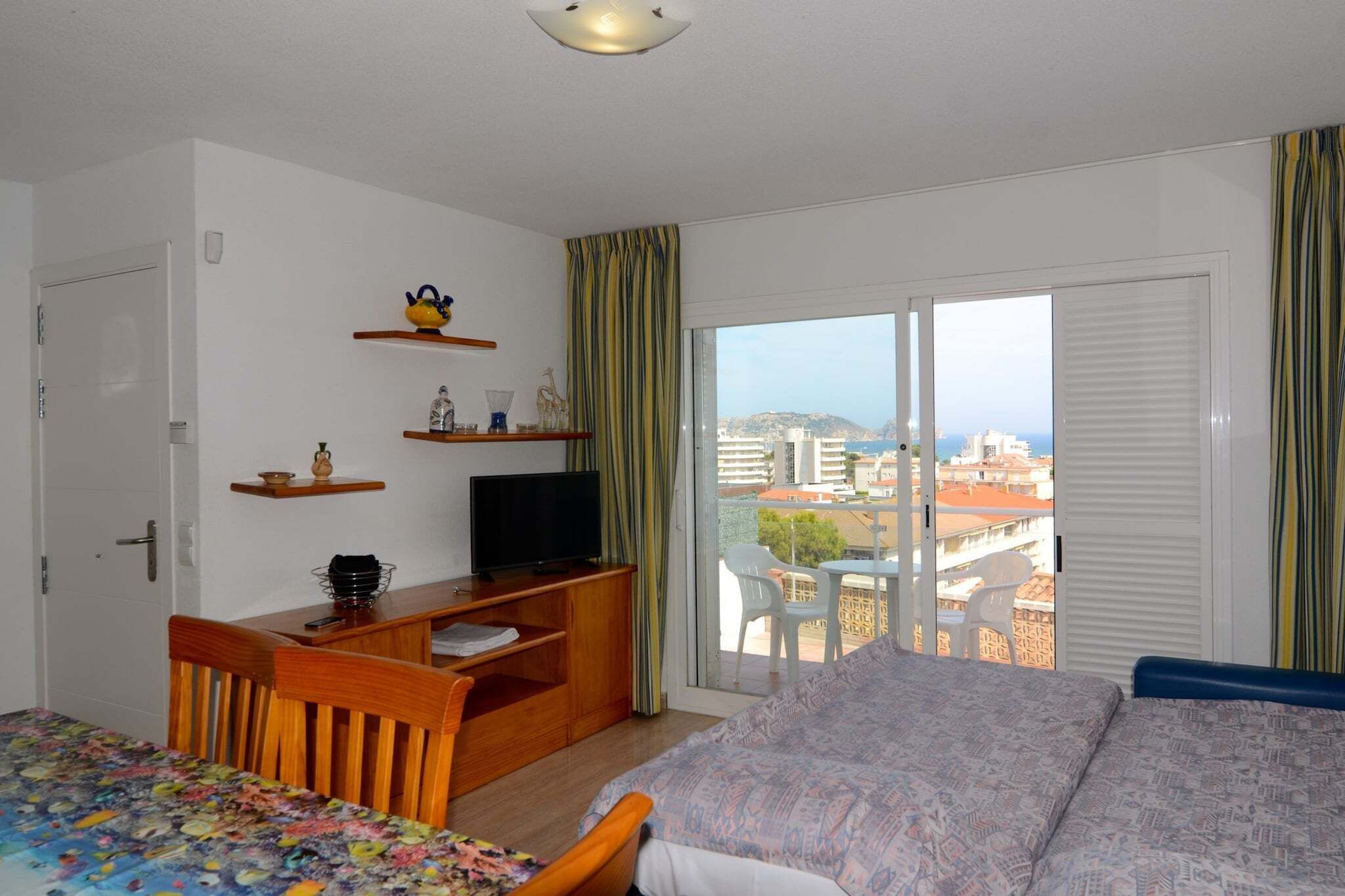 A hotel room with a bed, a desk with a television, a chair, a small table, and a balcony with a view of the city.