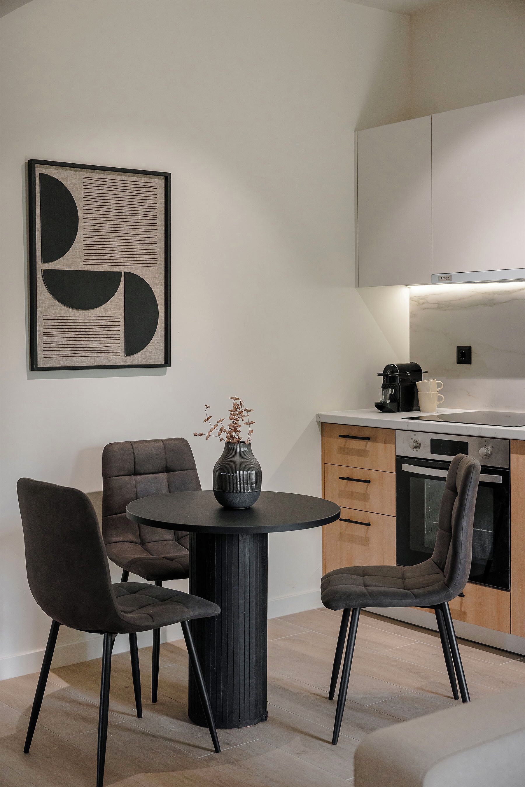 A modern kitchen with a dining area, featuring a round table, two chairs, a vase with dried flowers, and a coffee maker.