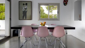 A modern dining area with a long table, pink chairs, a framed picture, a window with a view of trees, and a decorative mask on the wall. - Villa Bacio - My Rental Homes (San Jose)