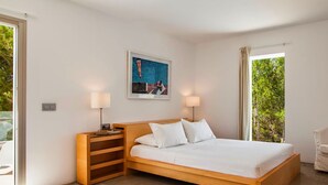 A modern bedroom with a wooden bed, two bedside lamps, a framed picture, and a view of greenery through a window. - Villa Bacio - My Rental Homes (San Jose)