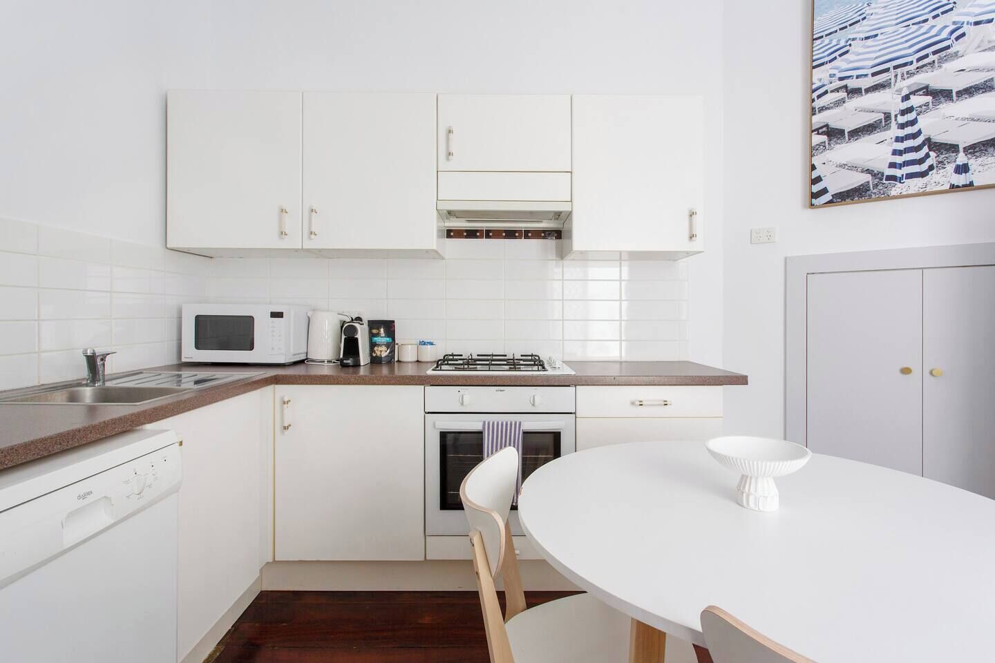A compact kitchen with white cabinets, a microwave, a coffee maker, and a stove. There is a white dining table with chairs.