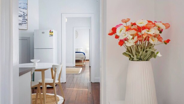 Une chambre d’hôtel moderne avec un plancher en bois, une table à manger blanche avec des chaises, un réfrigérateur blanc et un lit recouvert d’une couverture bleue.