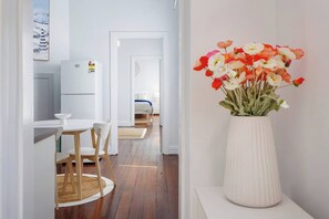 A modern hotel room with a wooden floor, a white dining table with chairs, a white refrigerator, and a bed with a blue blanket.