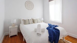 Une chambre à coucher comprenant un lit, une table de chevet, une lampe, une fenêtre avec des rideaux et un élément décoratif mural.