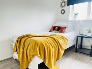 Uma cama com um cobertor amarelo-mostarda, roupa de cama branca e uma única almofada com um desenho de rosto. Uma pequena mesa com uma planta e um objeto decorativo fica ao lado da cama.