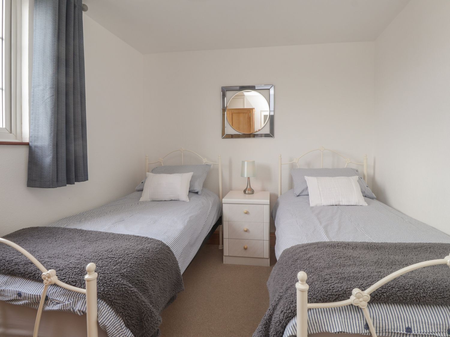 Un dormitorio con dos camas individuales, una mesita de noche con lámpara, un espejo y una ventana con cortinas.