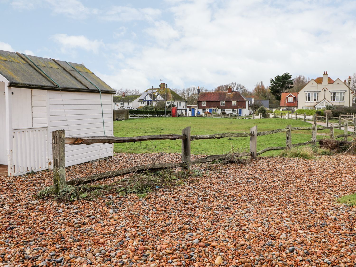 A gravel driveway leading to a white building with a metal roof, surrounded by a wooden fence and houses in the background.