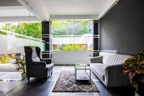 A modern living room with a sofa, armchair, and coffee table, a large window with a view of greenery, and a potted plant. - Housr 84 Kondapur Near Botanical Garden (Hyderabad)