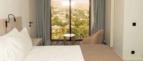 A hotel room with a large bed, a small table, and a view of a town through a large window.