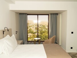 Habitación de hotel con una cama grande, una mesita pequeña y vistas de un pueblo a través de un ventanal amplio.