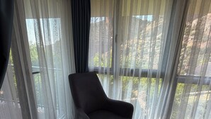 Una stanza con una poltrona scura vicino a una finestra con tende leggere, pavimento in legno e vista su un paesaggio naturale.