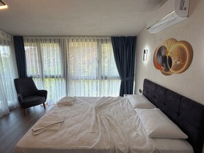 A hotel room with a large bed, a chair, a wall-mounted air conditioner, and a window with curtains.