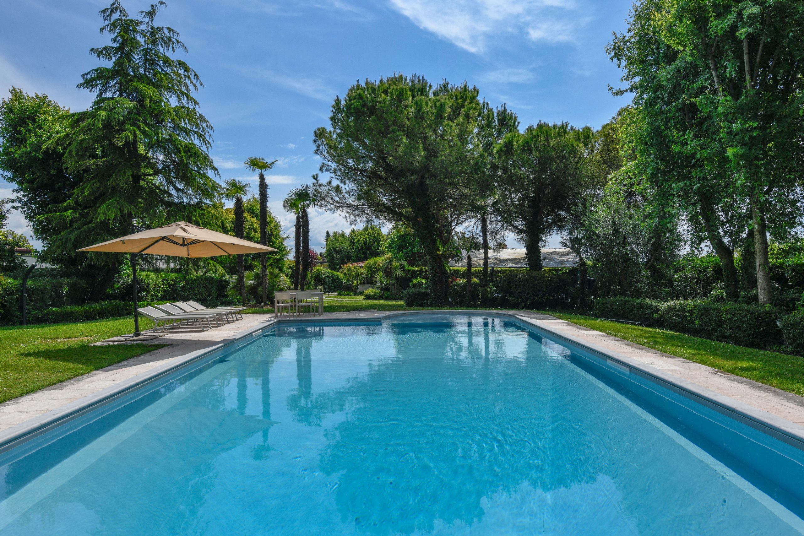 A rectangular swimming pool with a clear blue water surface, surrounded by lush green trees and a well-maintained lawn.