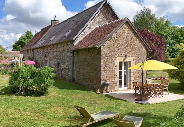 A stone building with a sloped roof, a patio with a table and chairs, and a small garden area.