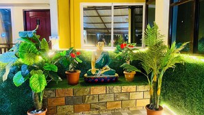 Une statue de Bouddha, entourée de plantes en pot et de fleurs, est placée sur une corniche en pierre devant un bâtiment jaune.