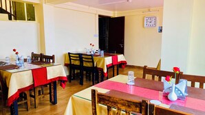 Un espace repas avec des tables préparées pour deux personnes, agrémenté de nappes rouges et blanches, de chaises en bois et d’une horloge au mur.