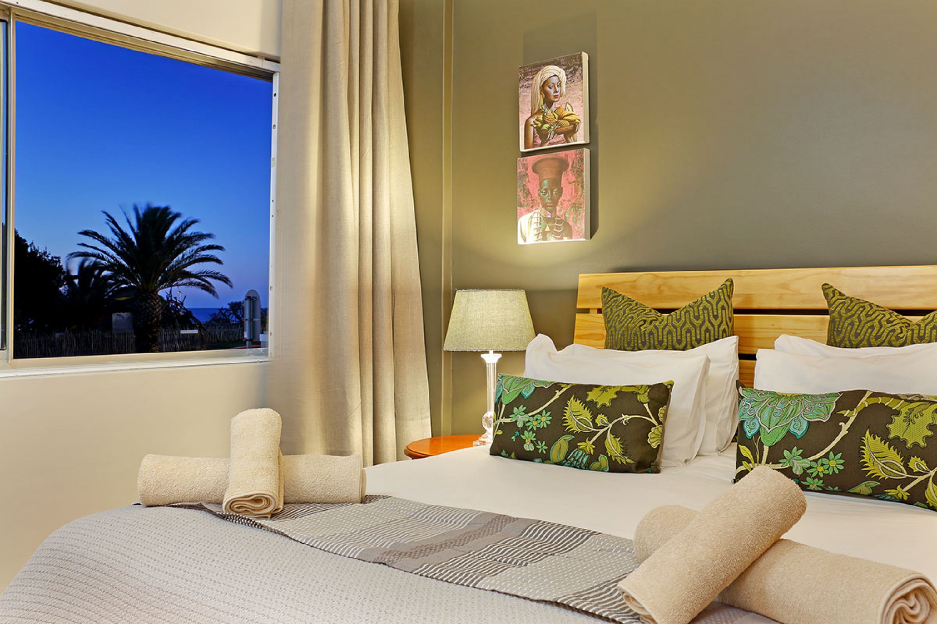 Una camera d'albergo con un letto, cuscini, una lampada, una finestra con tende e opere d'arte alle pareti.