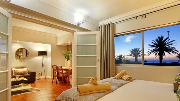 Un dormitorio con una cama grande, vistas a palmeras y una zona de comedor con una silla y una mesa.