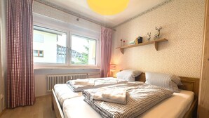 Une chambre avec deux lits, une fenêtre, un radiateur, une étagère avec des objets décoratifs et une lampe.