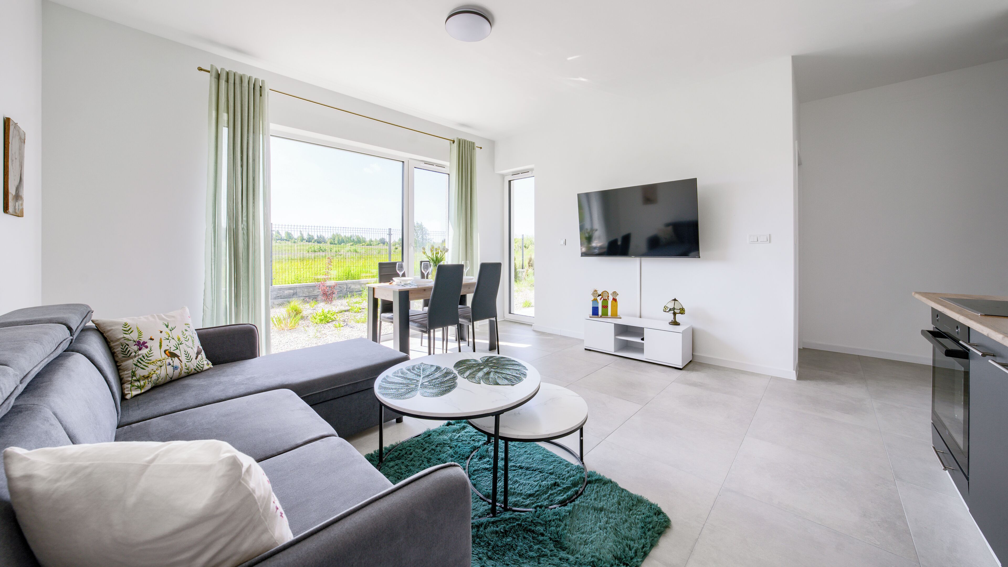 A modern living room with a grey sofa, a round coffee table, and a dining area with chairs and a table. A flat-screen TV is mounted on the wall, and there is a window with curtains.