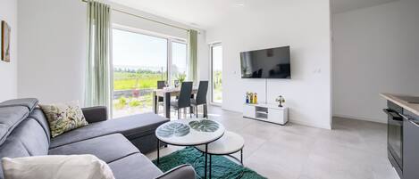 Ein modernes Wohnzimmer mit grauem Sofa, rundem Couchtisch und Essbereich mit Stühlen und Tisch. Ein Flachbildfernseher ist an der Wand befestigt, und es gibt ein Fenster mit Vorhängen.