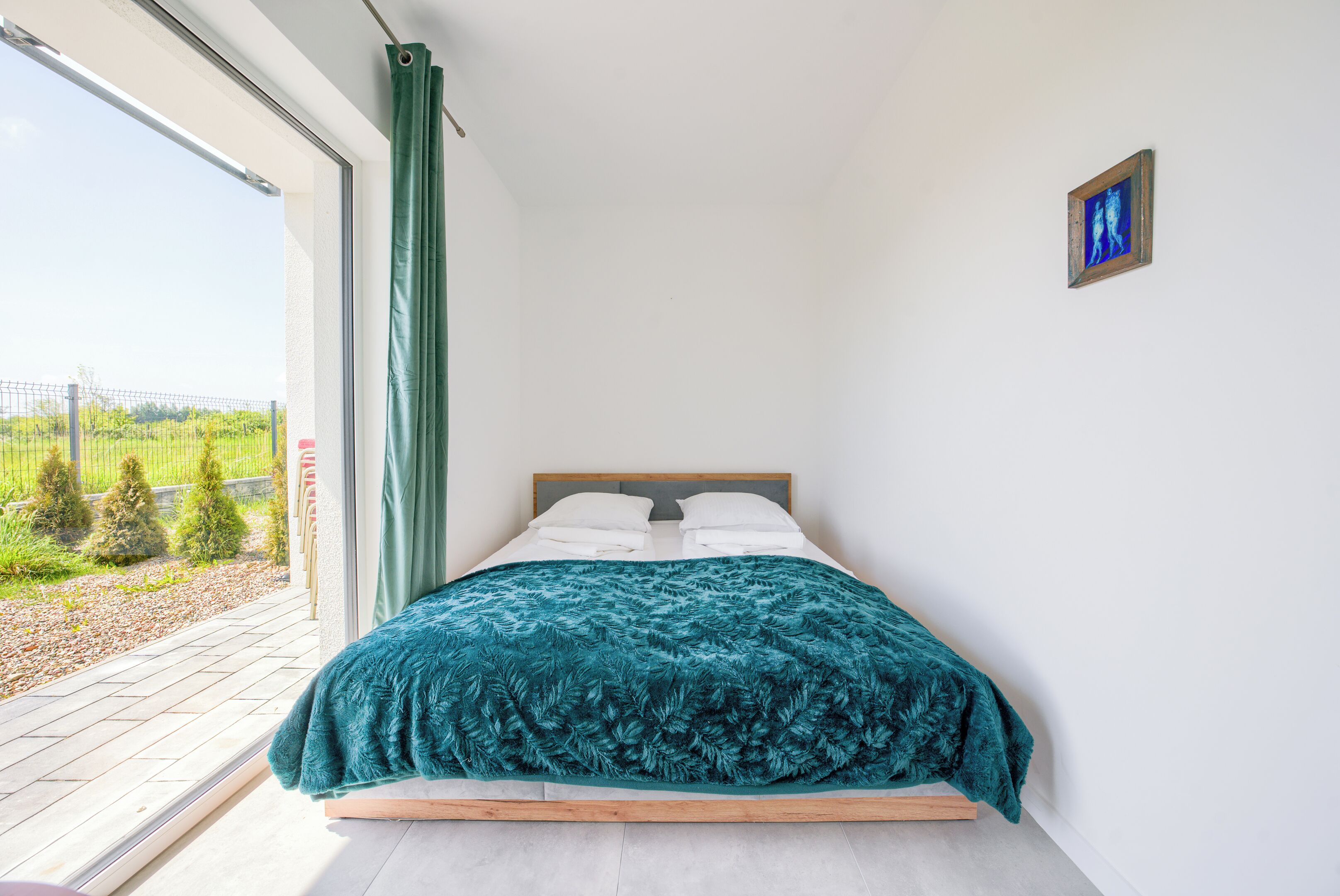 A bedroom with a bed, a window with a view of greenery, and a small picture on the wall.