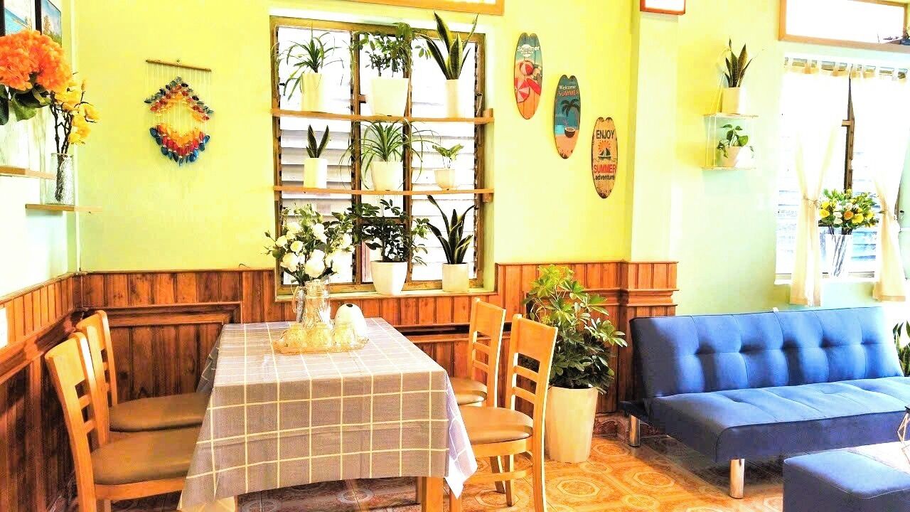 A dining area with a table set for two, wooden chairs, a blue sofa, and a window with potted plants.
