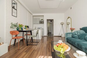 Uma sala de estar moderna com um sofá verde-água, uma área de jantar com cadeiras laranja e uma cozinha ao fundo.